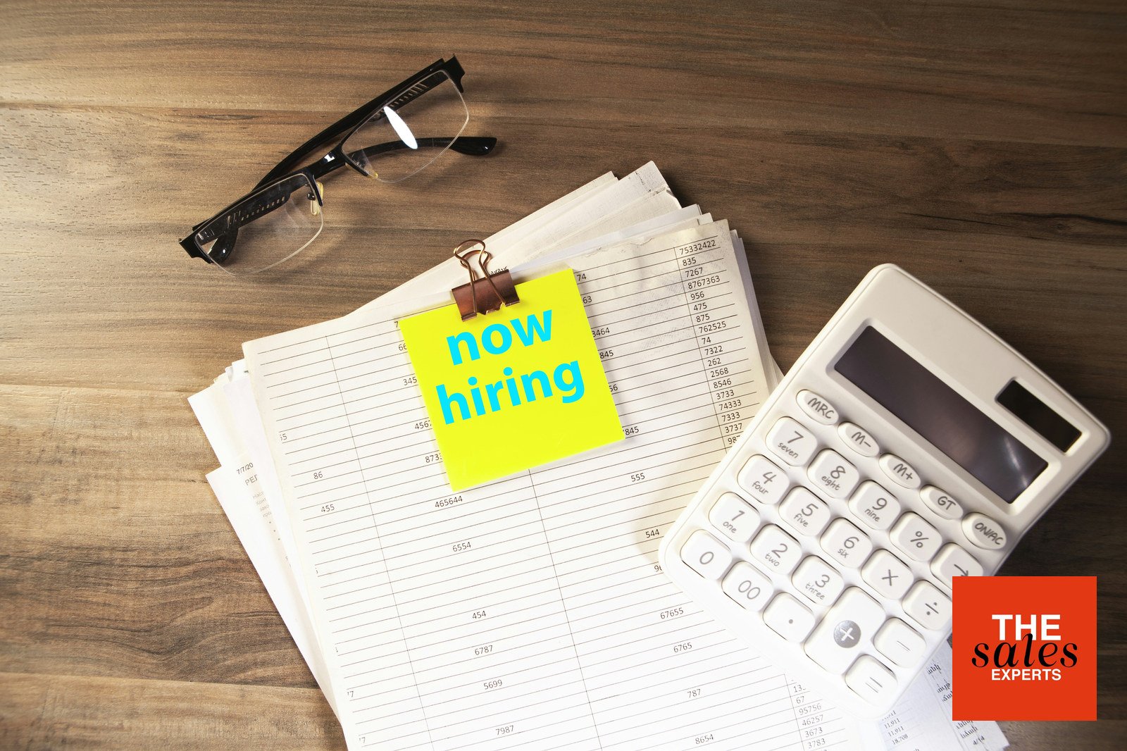 Now hiring note beside a calculator while planning the first sales hire